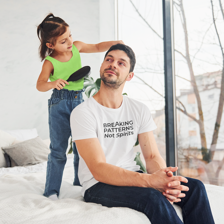 Young girl brushing her dad’s hair while he smiles, wearing a white t-shirt that says “Breaking Patterns Not Spirits” – gentle parenting Father’s Day shirt by The Unfussy Thread Collective.