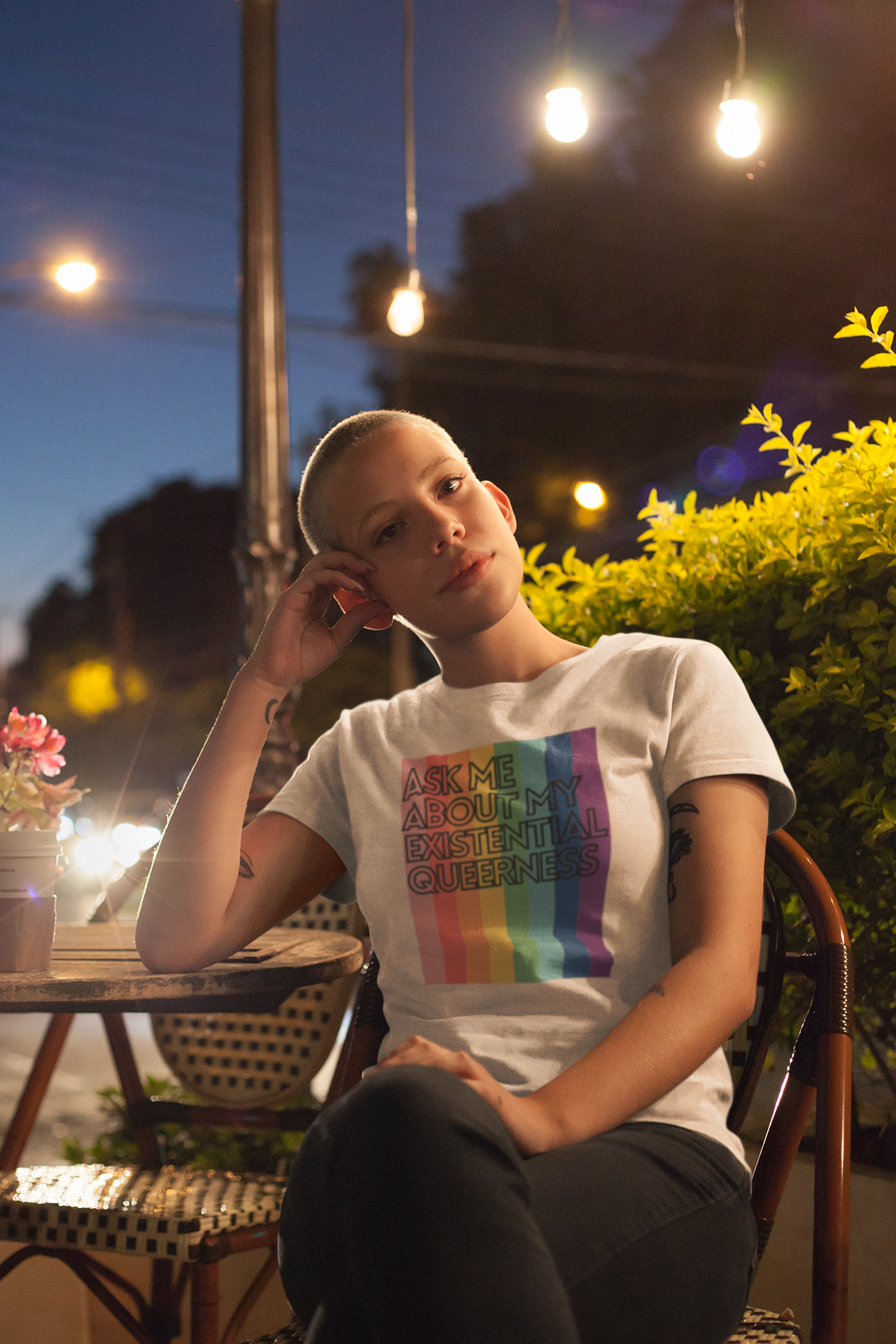 A person with short hair sits thoughtfully at an outdoor café at night, under glowing lights. They wear a white t-shirt with a rainbow-striped background and the words “Ask Me About My Existential Queerness.” Their pose and expression are calm, introspective, and proud.