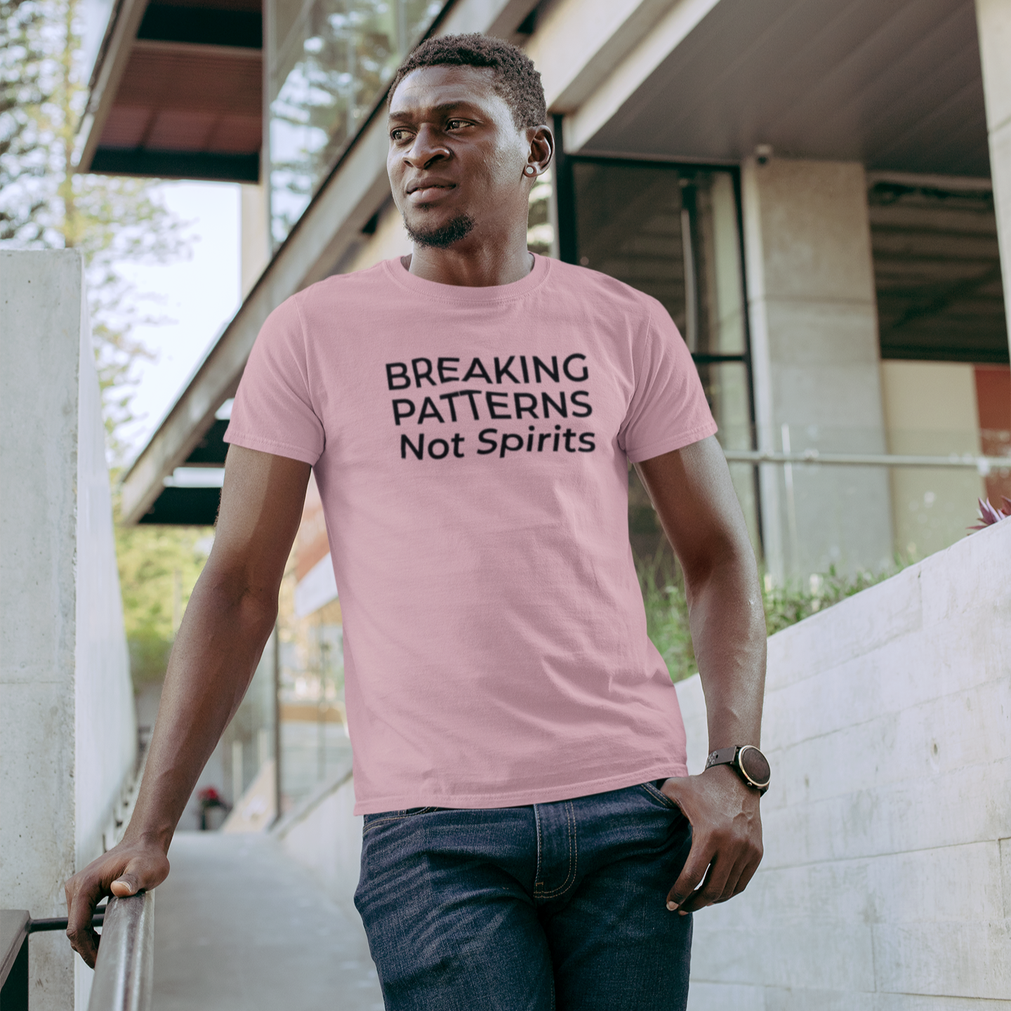 A man walks through a modern urban setting wearing a soft pink “Breaking Patterns Not Spirits” t-shirt by The Unfussy Thread Collective.