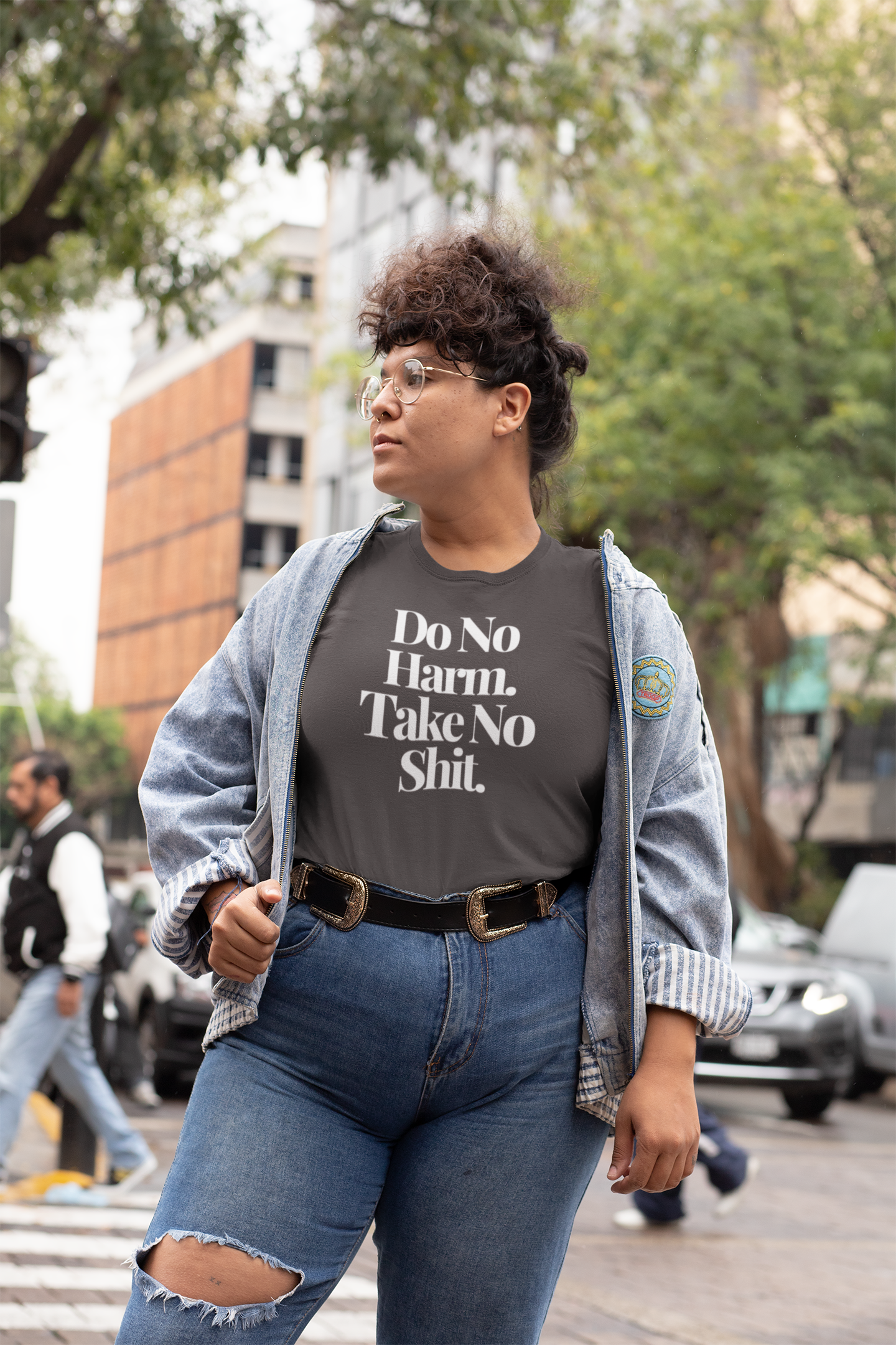Wearing glasses, a denim jacket, and The Unfussy Thread Collective's "Do No Harm Take No Shit" tee from The Plainspoken Collection in asphalt, someone strides confidently down a city street lined with trees and buildings—timeless Gen X style.