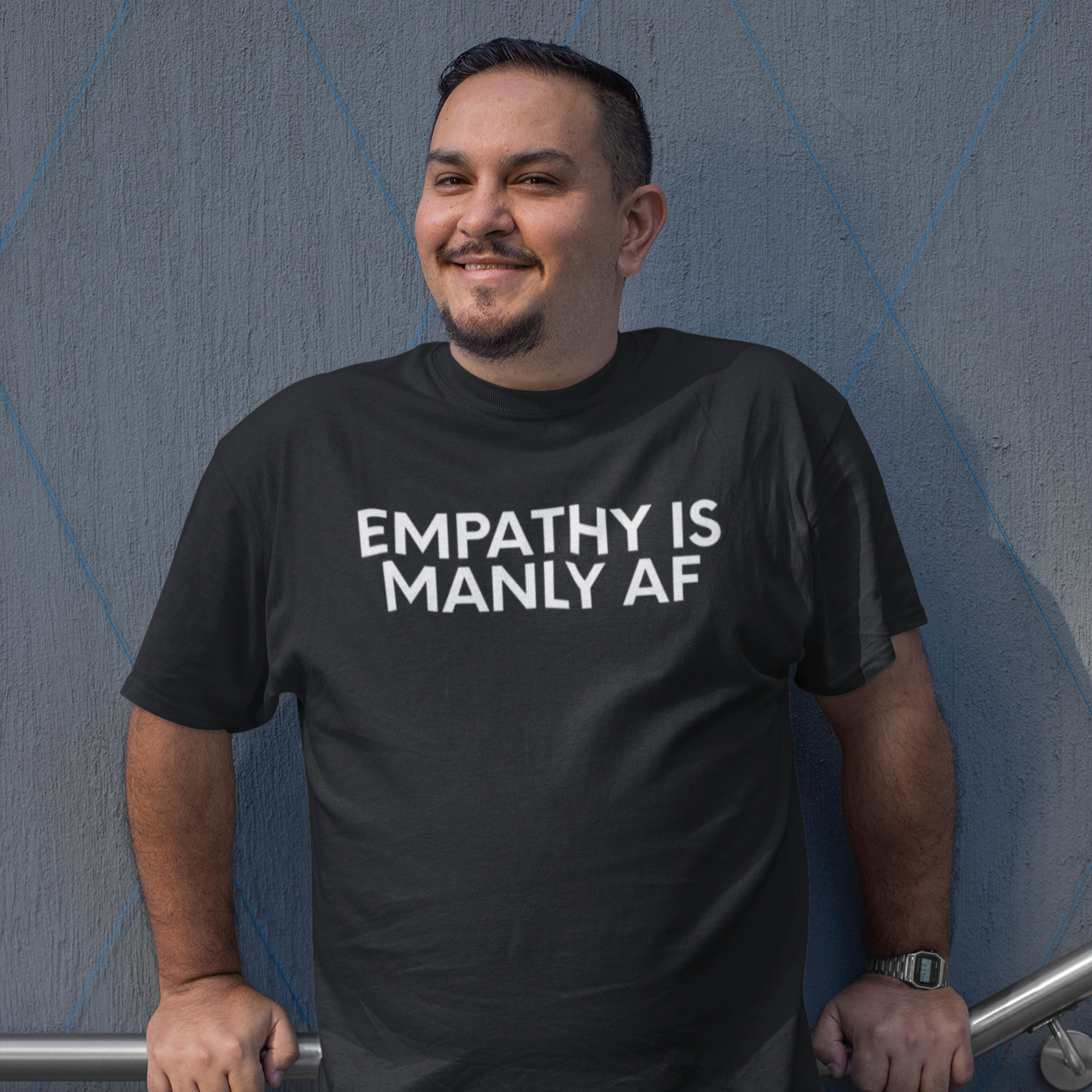 A smiling plus-size man leans casually against a wall wearing a Vintage Black “Empathy Is Manly AF” t-shirt from The Unfussy Thread Collective.