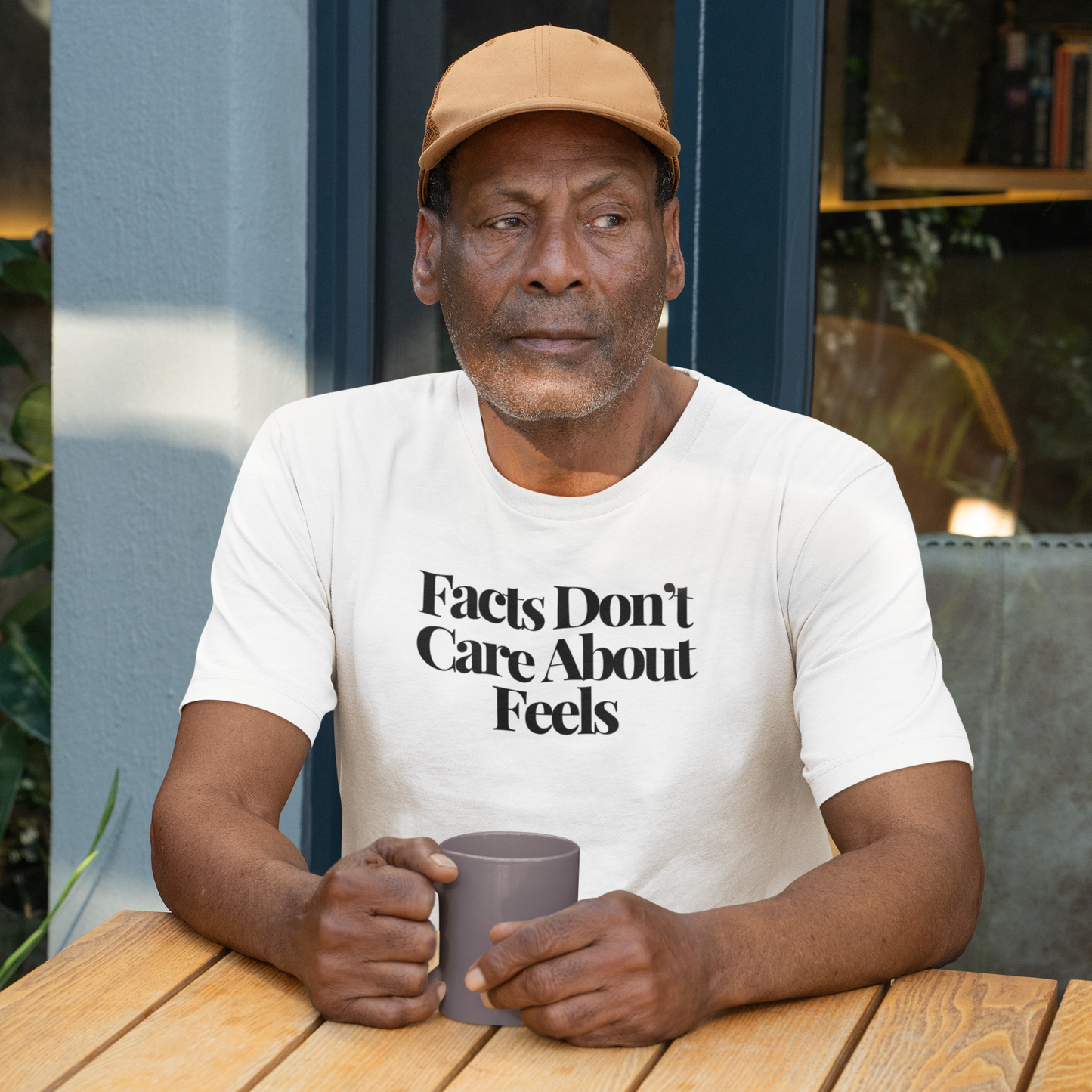 Older man wearing a tan cap and a white “Facts Don’t Care About Feels” t-shirt, sitting outdoors at a wooden café table, holding a gray mug with a thoughtful expression.