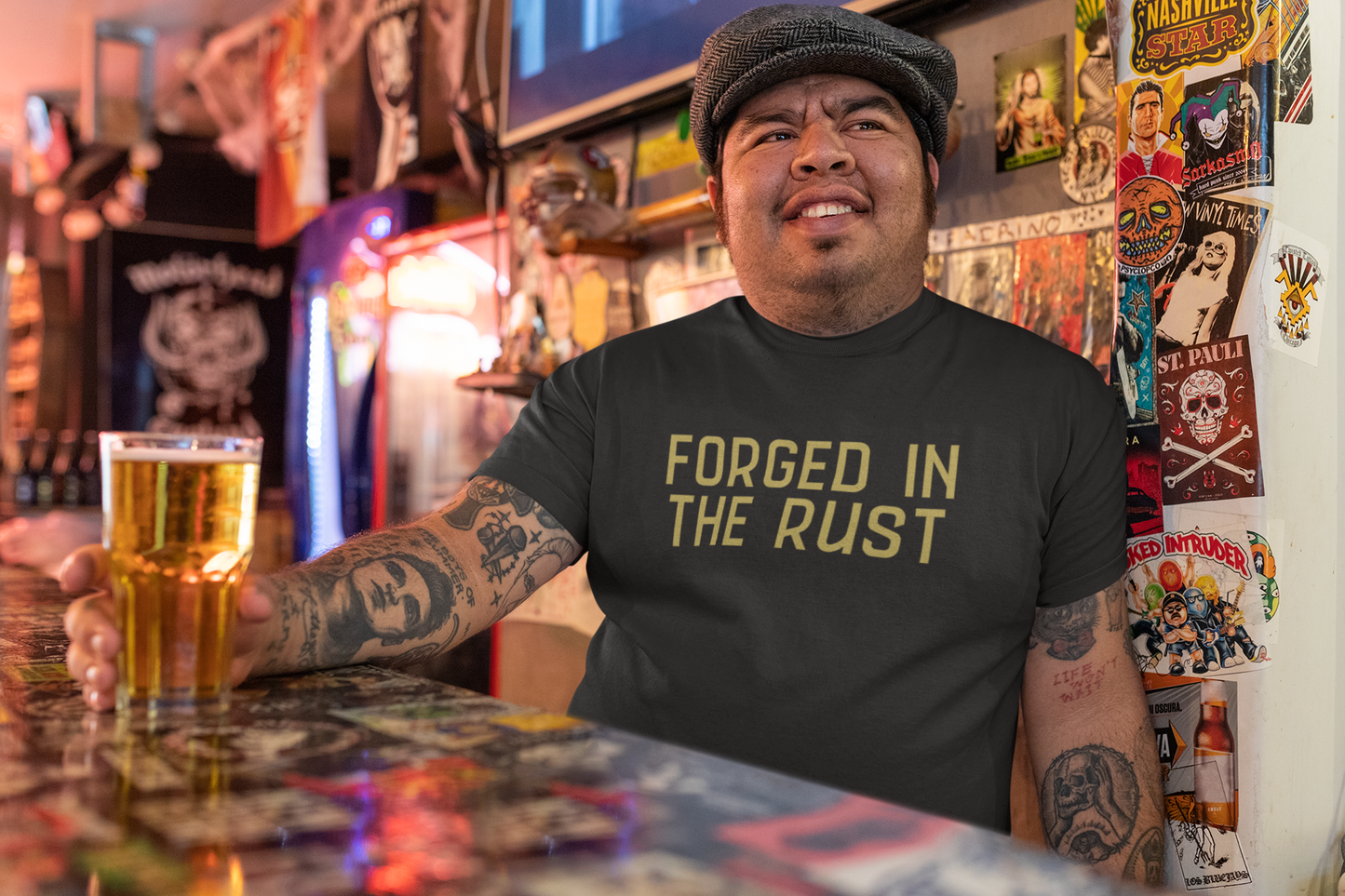 Tattooed man wearing a “Forged in the Rust” t-shirt while holding a beer at a bar, smiling confidently in a bold, punk-inspired setting.