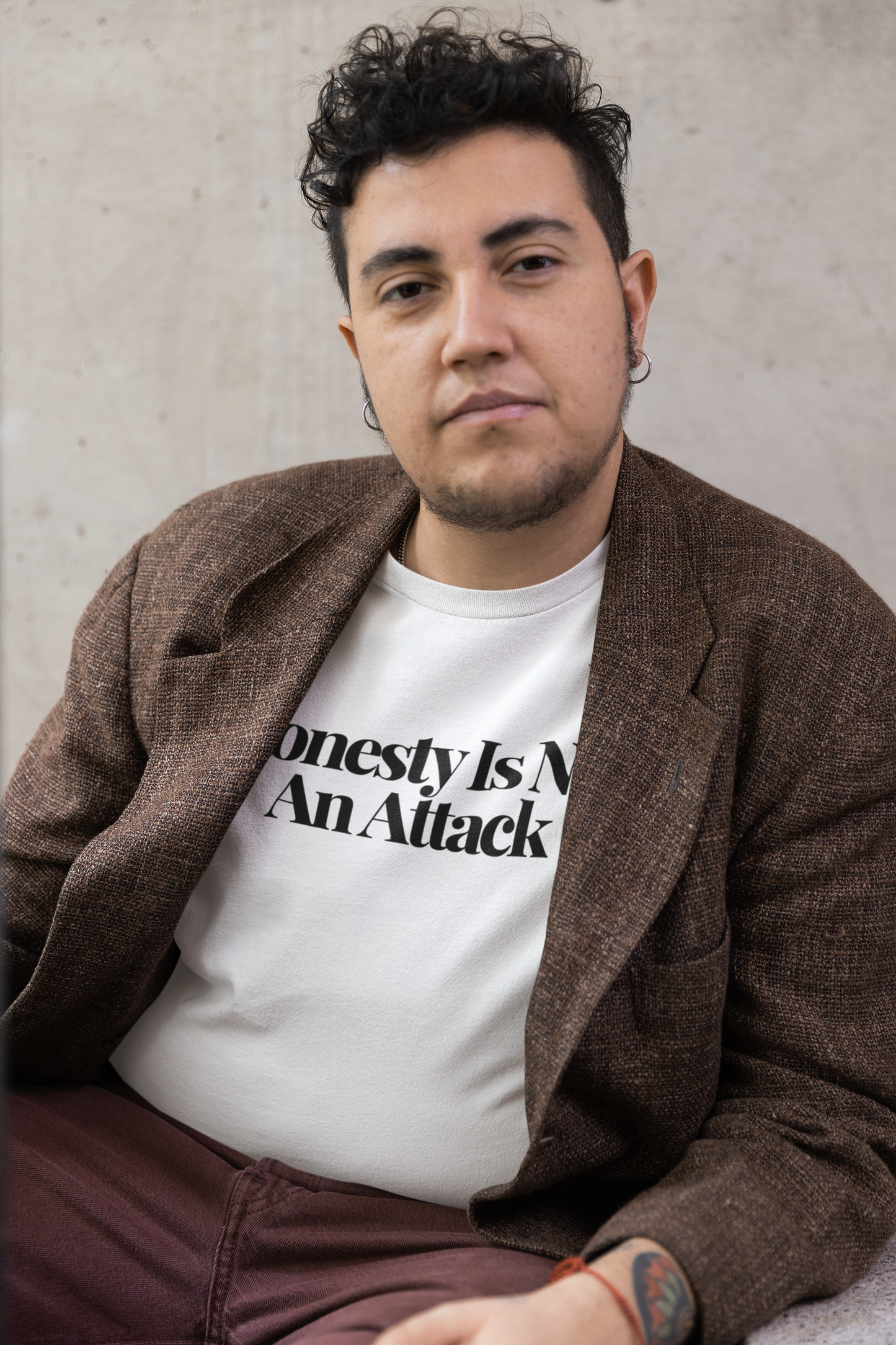 Person with short curly hair wearing a brown textured blazer over a white “Honesty Is Not An Attack” t-shirt, seated against a minimalist concrete wall.