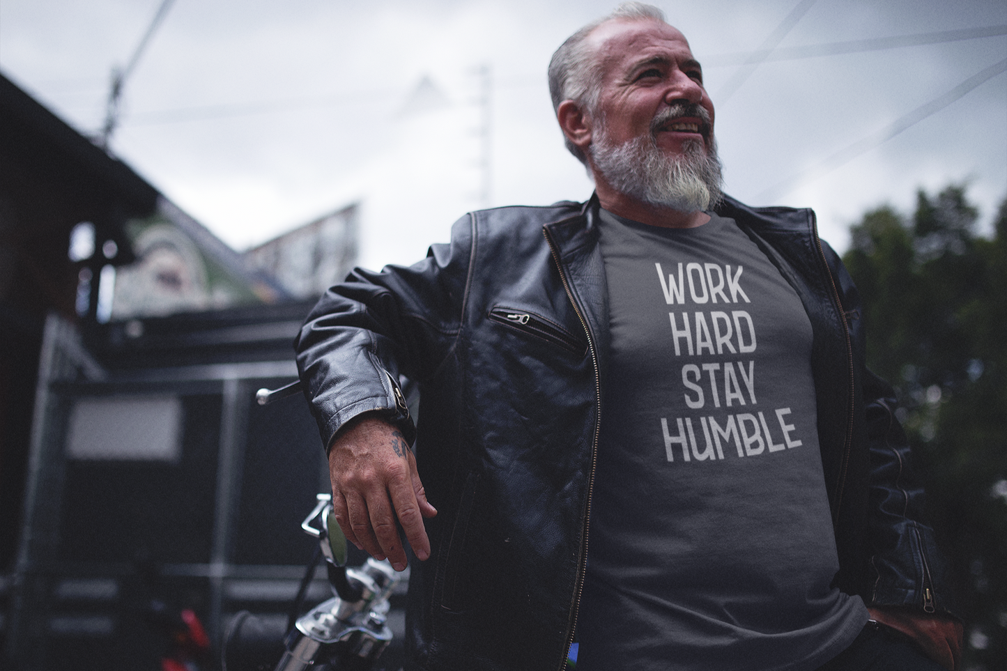 Older man in a leather jacket standing confidently next to a motorcycle, wearing a charcoal t-shirt that says "Work Hard Stay Humble" in bold lettering.