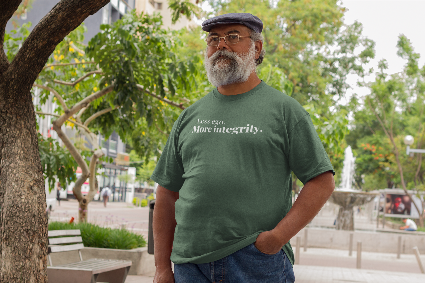 Older man with a gray beard and glasses standing outdoors near a fountain, wearing a green “Less ego. More integrity.” t-shirt and a newsboy cap.