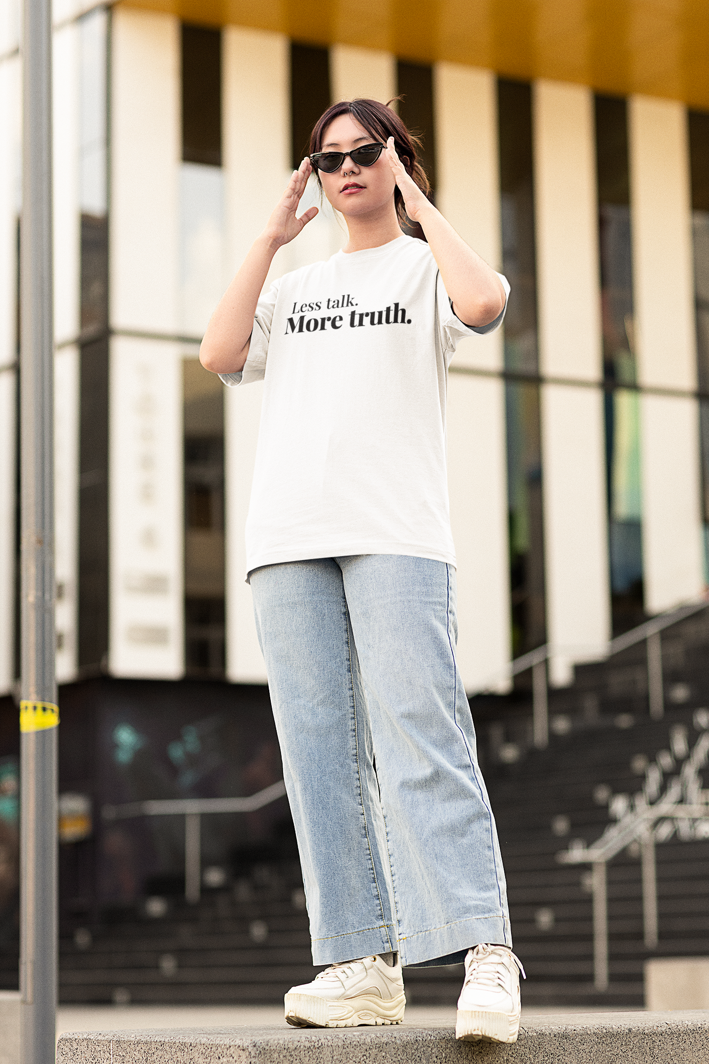 Trendy young woman wearing sunglasses and a white “Less talk. More truth.” oversized t-shirt, standing confidently outdoors in an urban area with a modern building in the background.