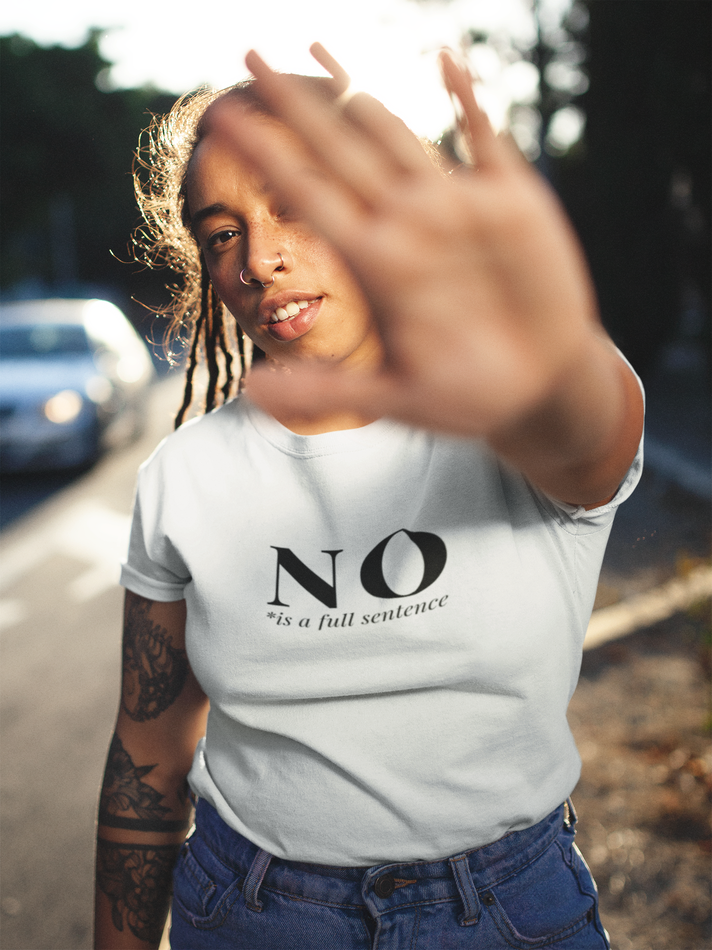 Confident young woman with tattoos and nose piercings wearing a white t-shirt that says “NO is a full sentence,” holding her hand up toward the camera in a powerful gesture while standing on a sunlit street.