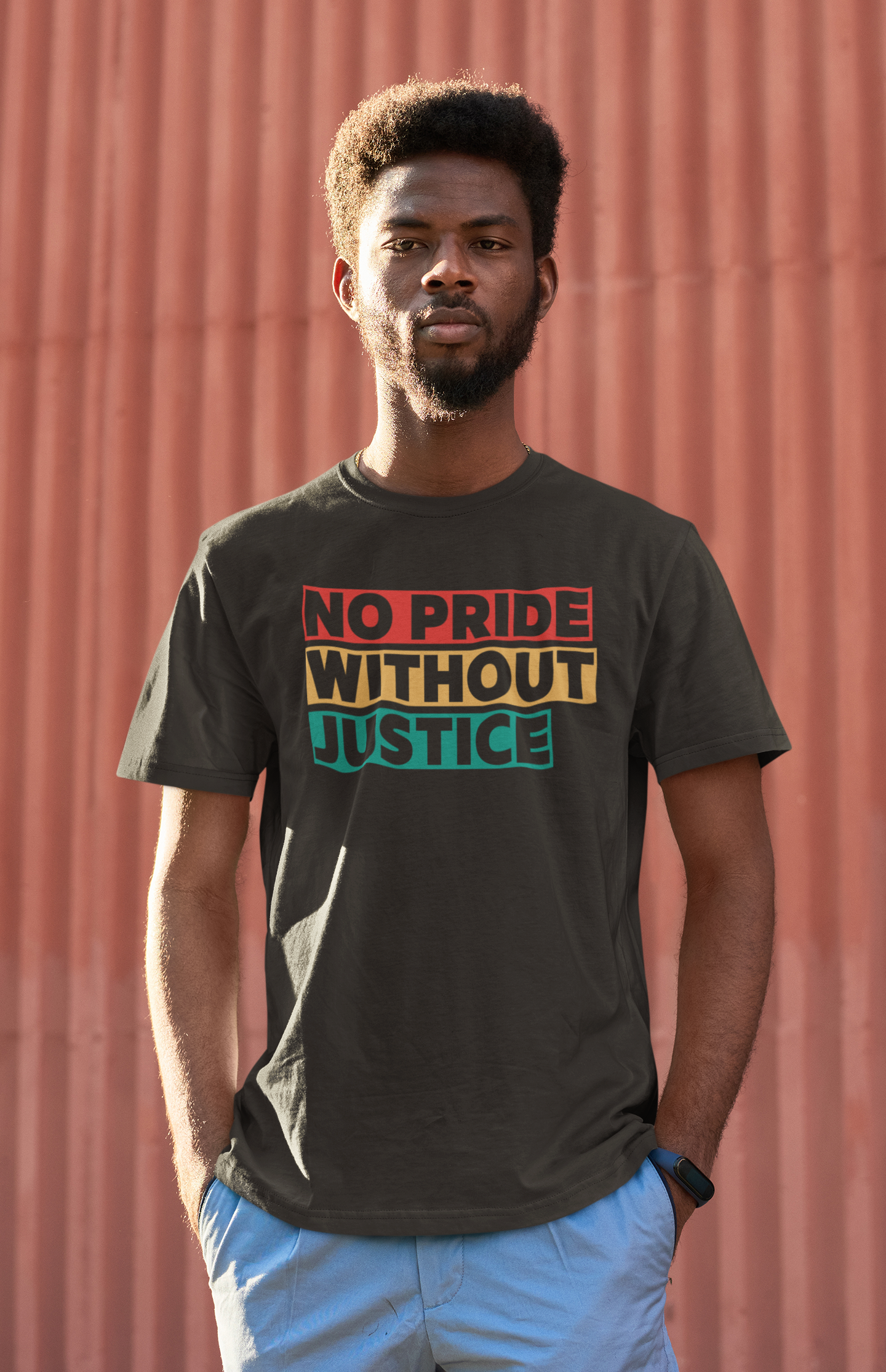 A serious young Black man stands in front of a textured, rust-red wall wearing a dark t-shirt that reads “No Pride Without Justice” in red, gold, and green block letters. His hands are in his pockets and the sunlight casts strong shadows, emphasizing the shirt’s powerful call for intersectional justice and racial equity within the LGBTQIA+ movement.