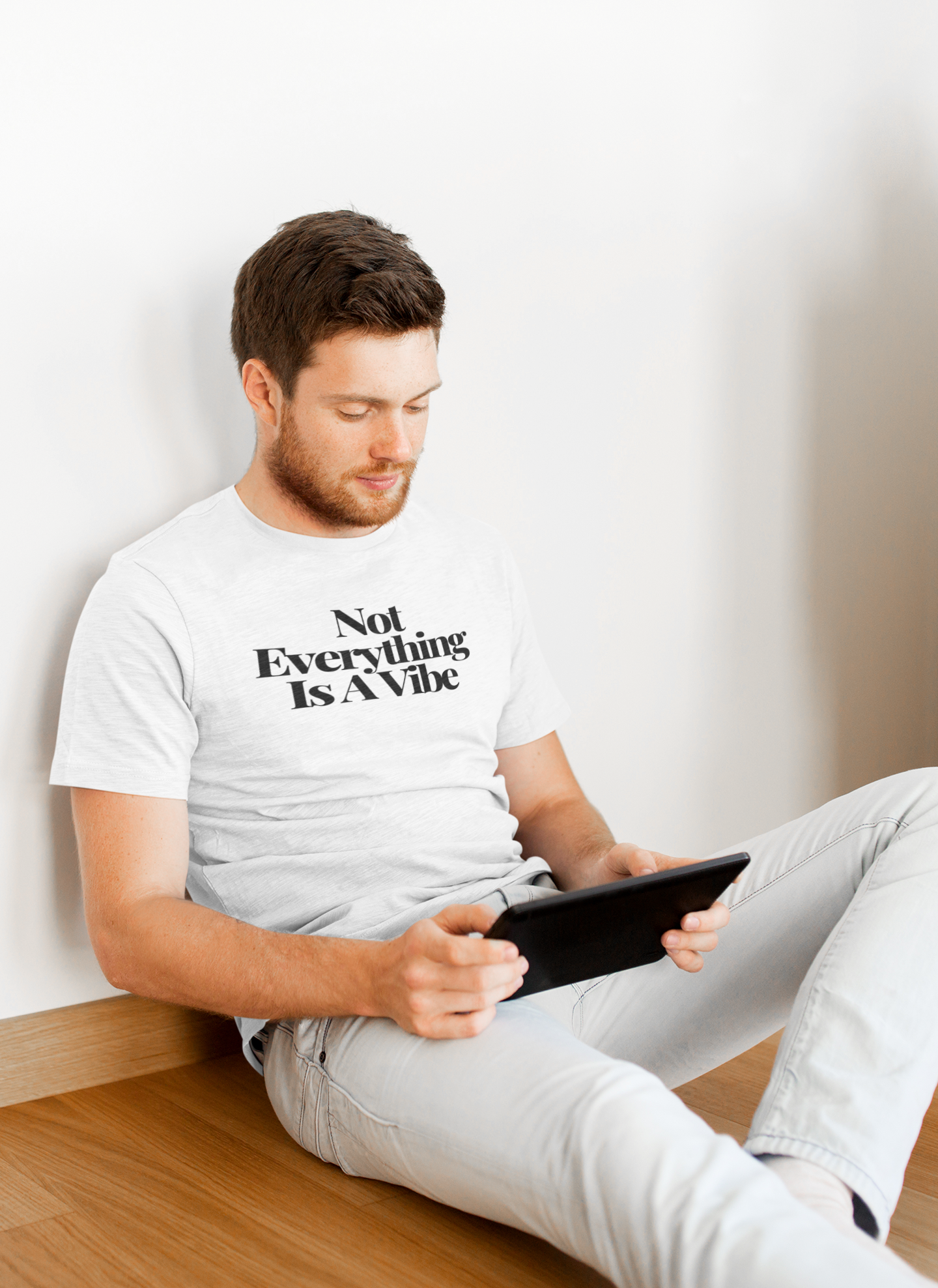 Young man with short brown hair and a beard sits casually on a wooden floor, leaning against a white wall, wearing a light “Not Everything Is A Vibe” t-shirt while using a tablet. The clean, neutral background highlights the minimalist, thoughtful vibe of the scene.