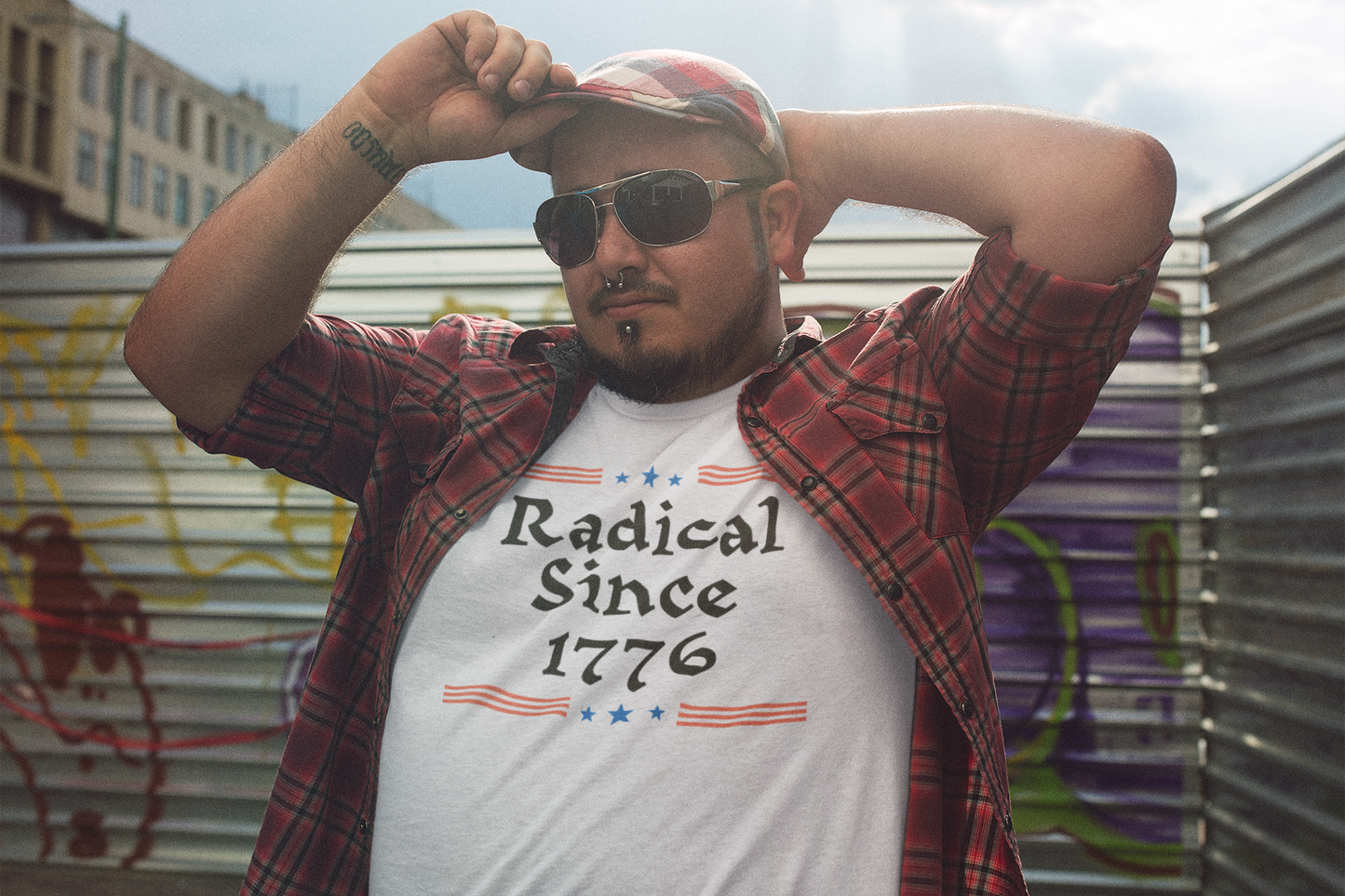 A man in sunglasses and a red flannel shirt poses confidently in an urban alleyway, wearing a white t-shirt that reads “Radical Since 1776” with red and blue graphics.