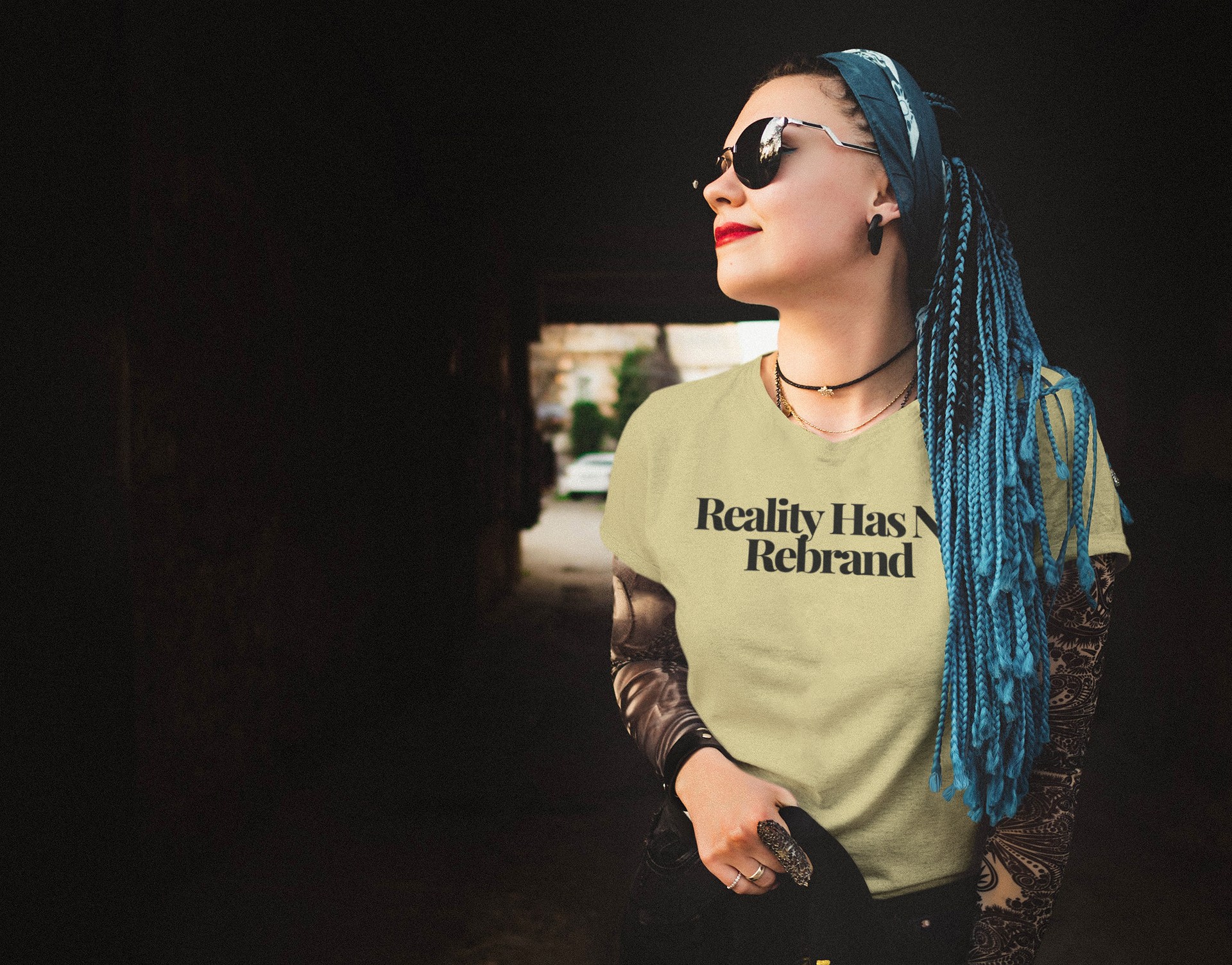 A smiling person with blue braided hair, sunglasses, and tattoos stands outside in The Unfussy Thread Collective’s "Reality Has No Rebrand" tee from the Plainspoken Collection, color Natural. They look confidently aside, one hand in their pocket.