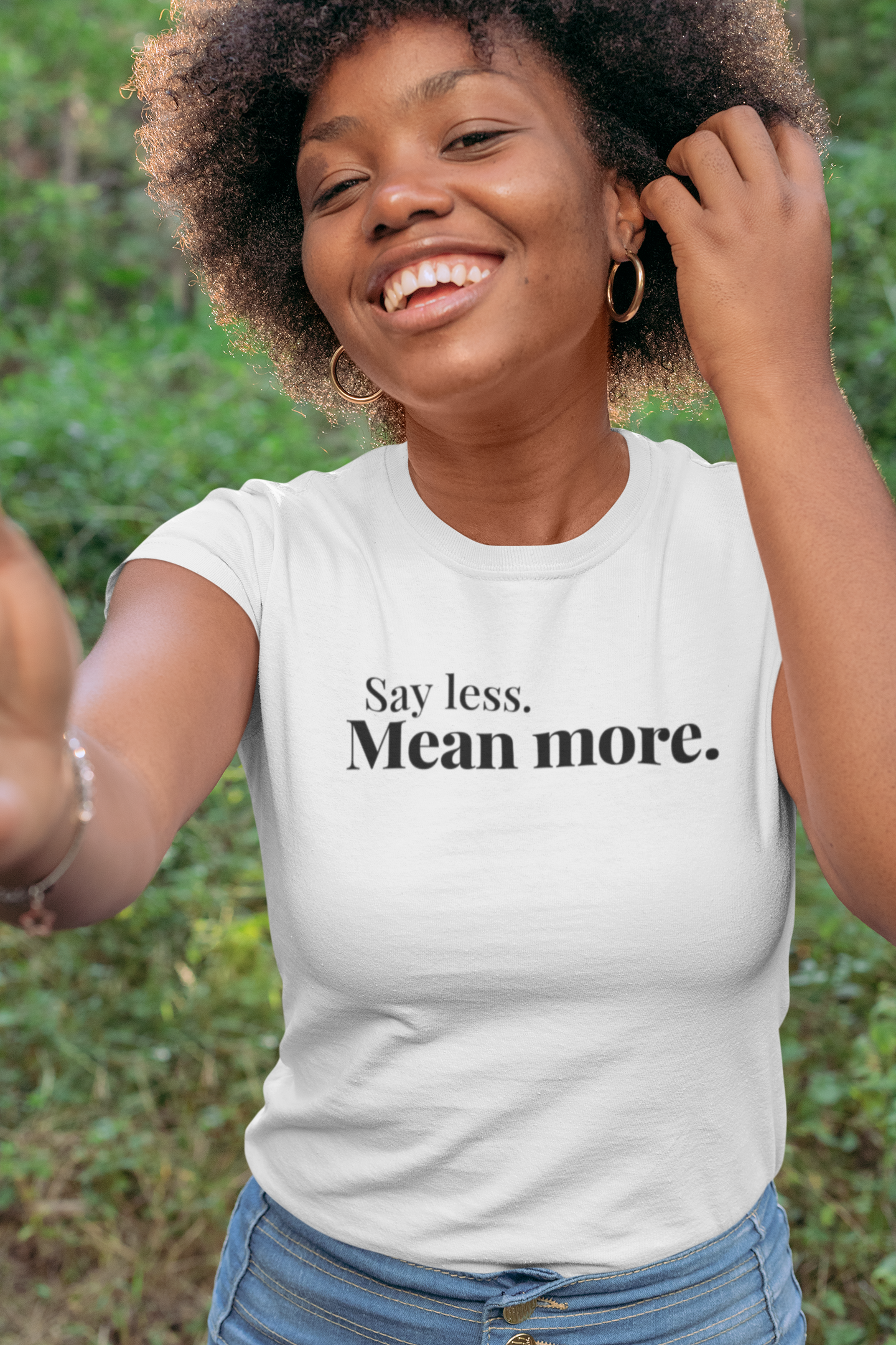 Smiling young woman with a natural afro hairstyle wearing a white “Say Less. Mean More.” t-shirt, standing outdoors surrounded by trees and greenery, exuding joy and confidence.