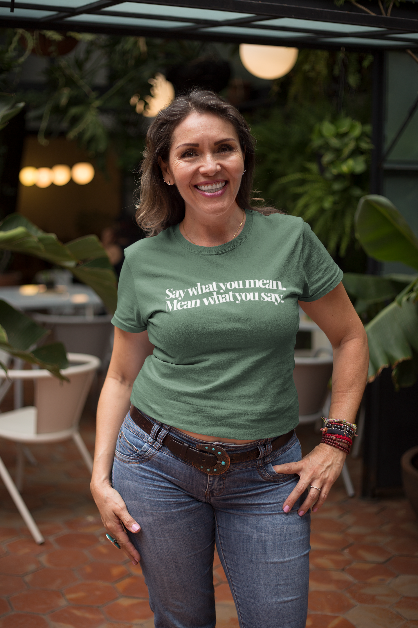 Confident smiling woman standing in a cozy coffee shop wearing a green “Say What You Mean. Mean What You Say.” t-shirt, styled casually with jeans and colorful bracelets, surrounded by plants and warm lights.
