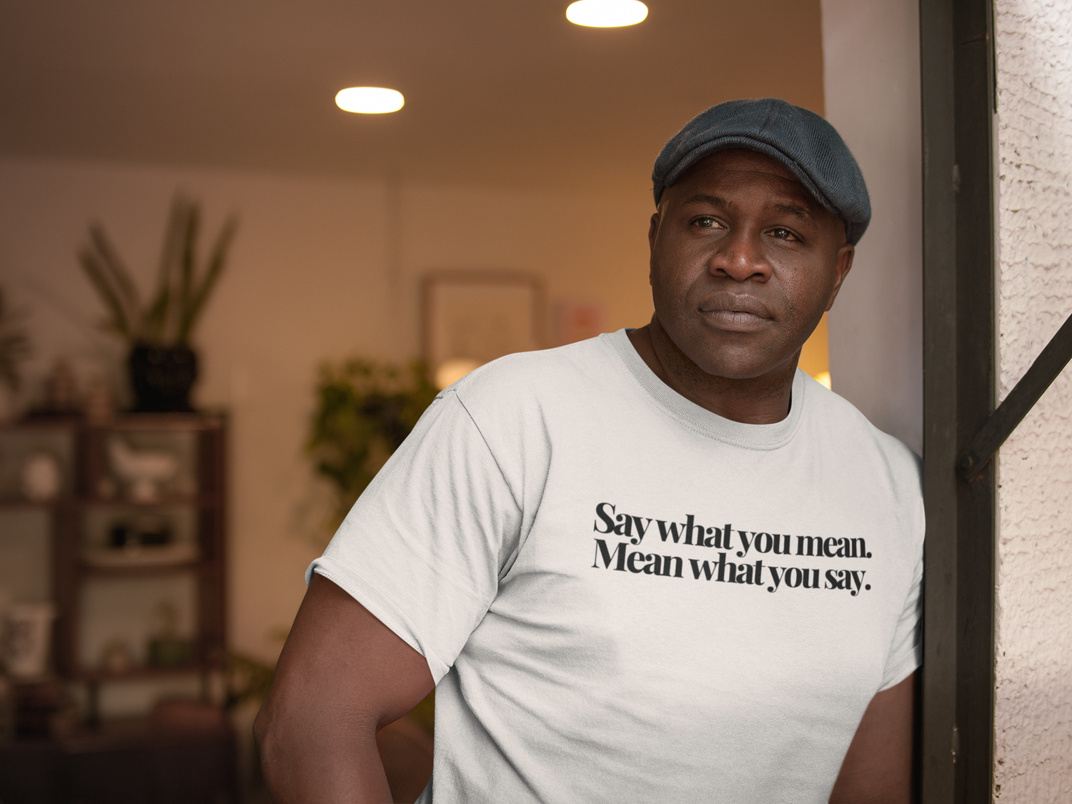 A man in a flat cap and Classic White "Say What You Mean, Mean What You Say" T-shirt from The Plainspoken Collection by The Unfussy Thread Collective stands by a doorway, gazing into a warmly lit room with plants and shelves.