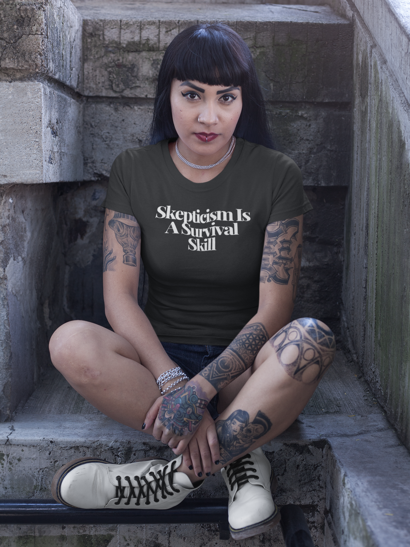 Tattooed woman with dark hair and bangs sits cross-legged on a stone stairway, wearing a dark “Skepticism Is A Survival Skill” t-shirt. Her intense gaze and bold style amplify the shirt’s message about resilience and critical thinking.