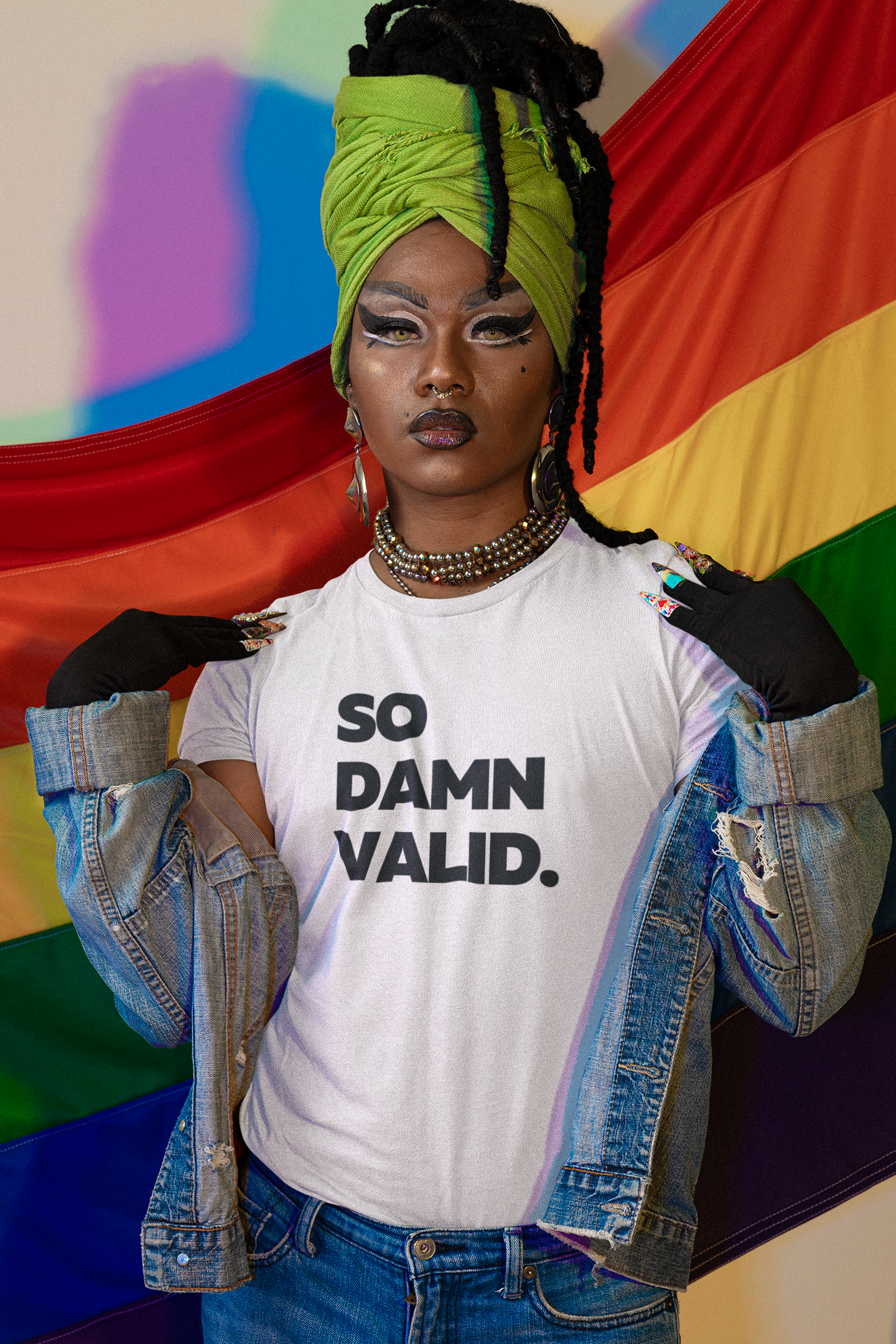 A person in a green headwrap, bold makeup, denim jacket, and The Unfussy Thread Collective's "So Damn Valid – Everyday Affirmation Tee" in classic white confidently poses before a rainbow pride flag.
