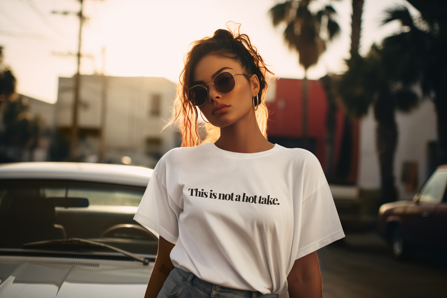 Stylish young woman wearing sunglasses and a white t-shirt that reads “This is not a hot take,” standing in front of a vintage car at sunset with palm trees in the background.