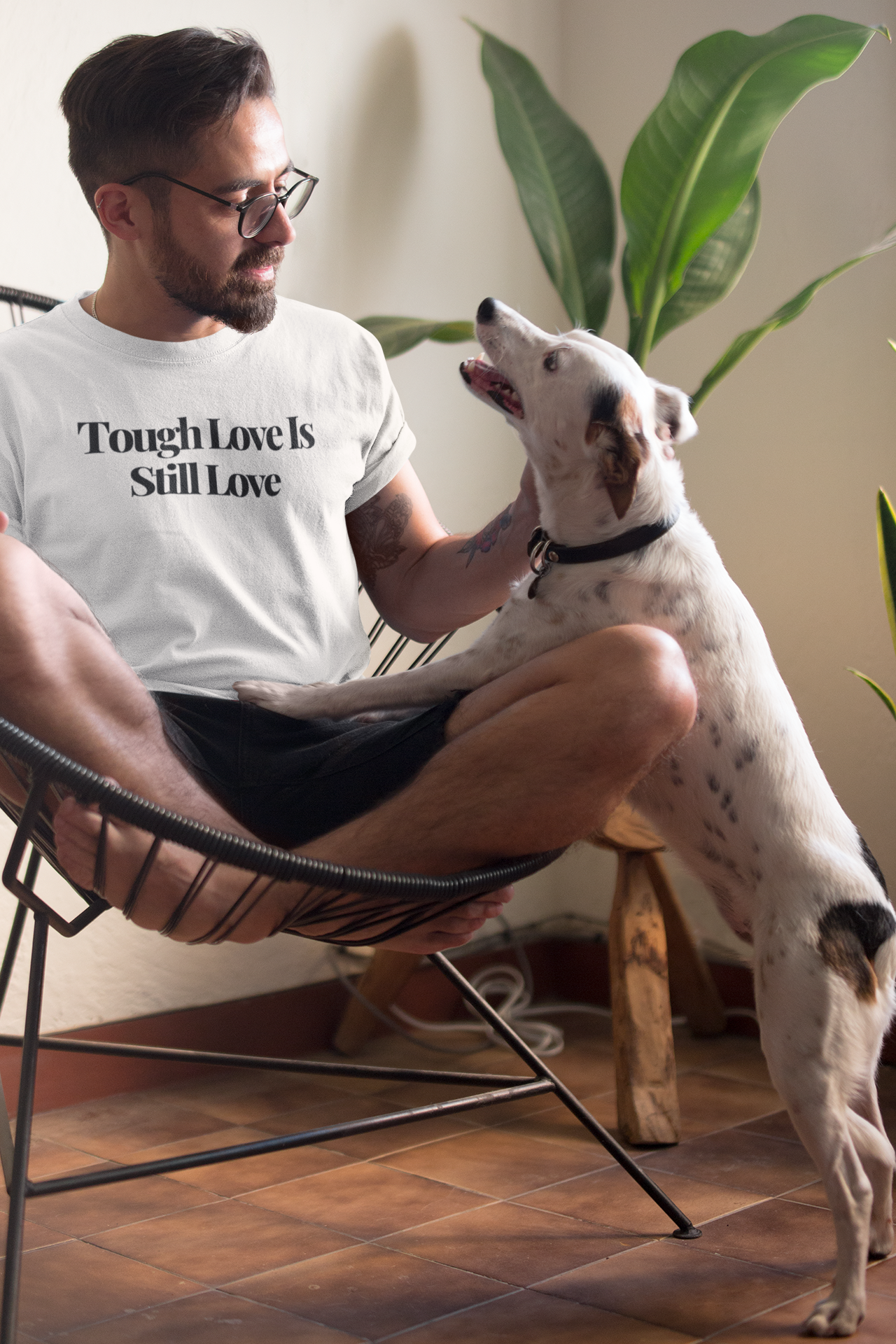 Bearded man sitting on a modern chair at home wearing a white “Tough Love Is Still Love” t-shirt while smiling and playing with a happy dog.