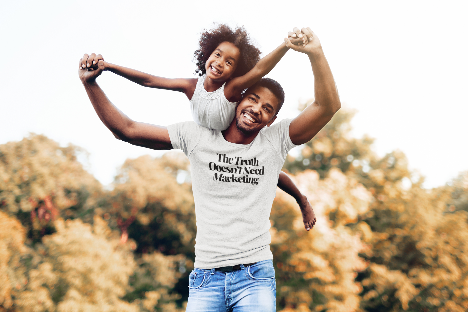 A smiling man lifts a joyful young girl onto his back outdoors, wearing The Unfussy Thread Collective’s Classic White “The Truth Doesn’t Need Marketing” tee from The Plainspoken Collection. Autumn trees blur in the background.