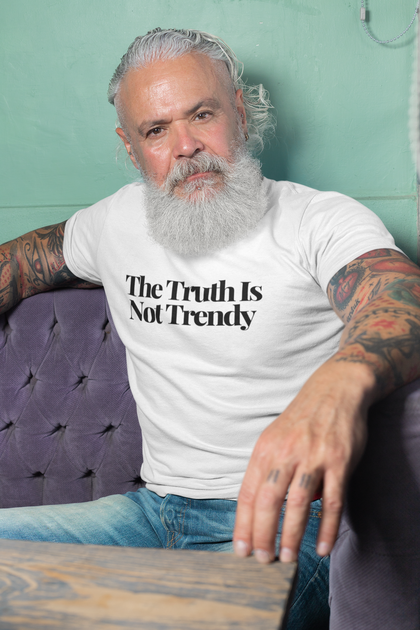 A bearded, tattooed man sits on a purple sofa, wearing a Classic White tee from The Unfussy Thread Collective’s So Damn Valid - The Plainspoken Collection. With a serious look, he embodies authentic style and the message “The Truth Is Not Trendy.”.