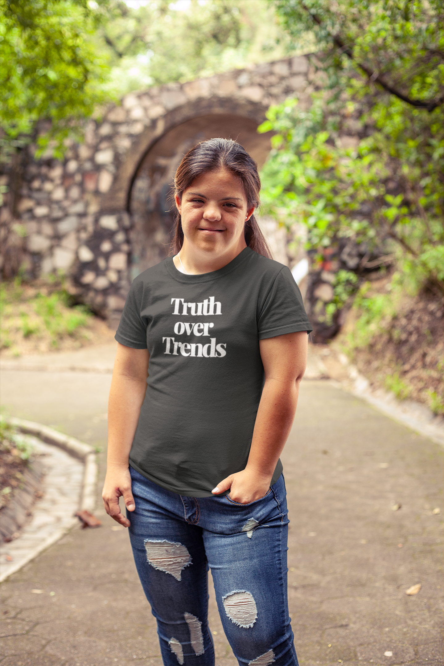Young woman with long brown hair smiles confidently while standing in a leafy park, wearing a dark “Truth Over Trends” t-shirt and distressed jeans. A rustic stone bridge arches in the background, adding to the natural, grounded atmosphere.