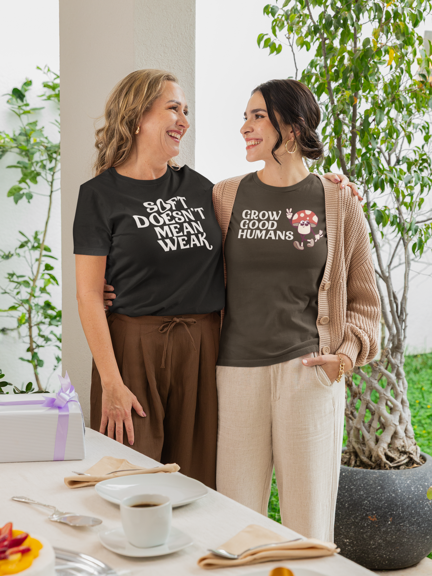 Two women smiling and hugging at a Mother’s Day celebration, wearing graphic t-shirts that say “Soft Doesn’t Mean Weak” and “Grow Good Humans” from The Unfussy Thread Collective’s This Is How We Mother collection.