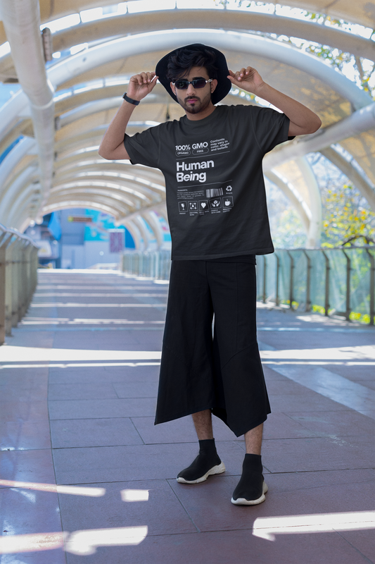 Person in black wide-leg pants and sunglasses wearing a dark t-shirt that reads "100% GMO-Free Organic Human Being" with white text styled like a nutrition label, standing confidently under a modern pedestrian bridge.