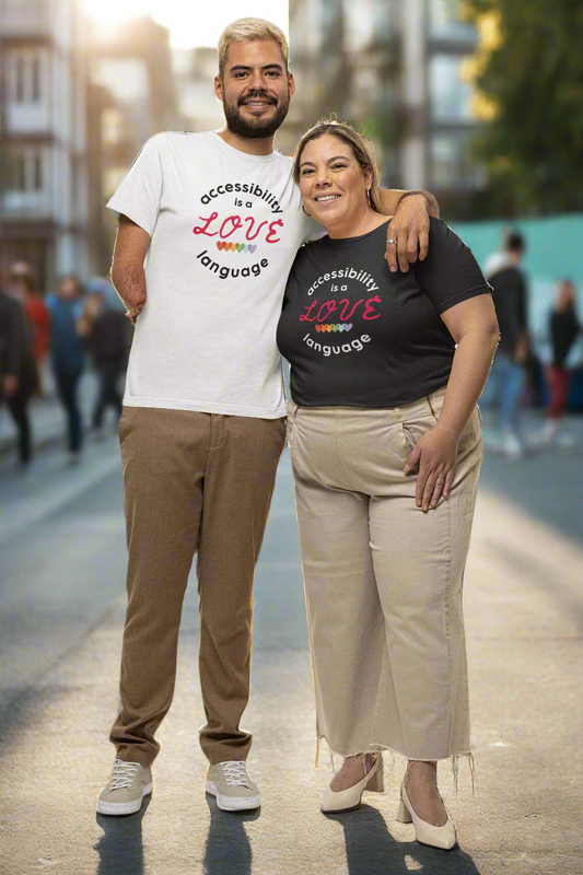 A smiling man and woman stand arm in arm outdoors, proudly wearing t-shirts that read “Accessibility is a Love Language.” The man wears a white tee and brown pants, and the woman wears a black tee and beige pants. Their warm expressions and confident stance radiate inclusion and pride.