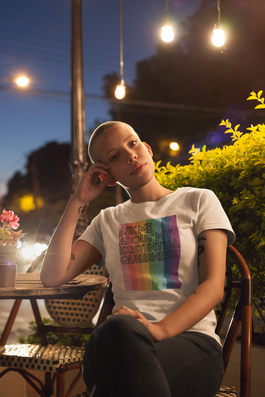 A person with short hair sits thoughtfully at an outdoor café at night, under glowing lights. They wear a white t-shirt with a rainbow-striped background and the words “Ask Me About My Existential Queerness.” Their pose and expression are calm, introspective, and proud.