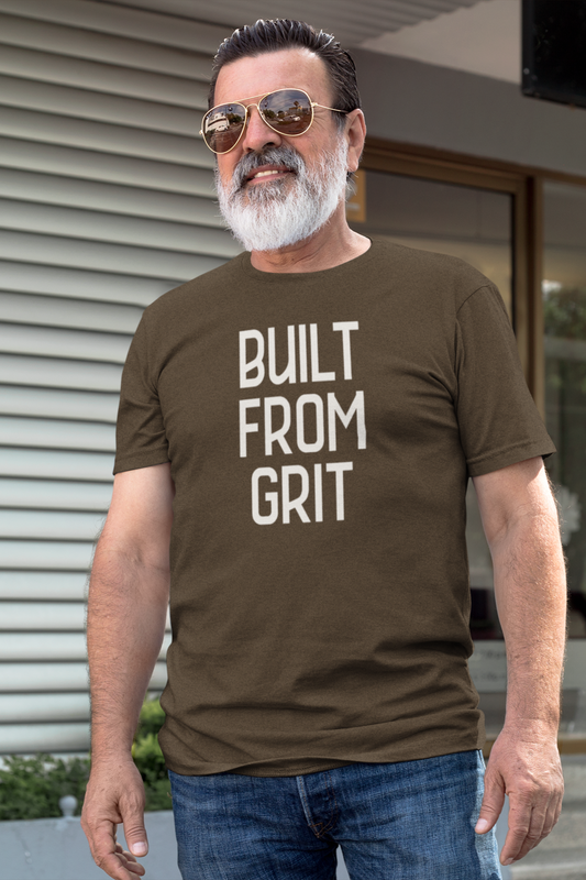 Older man with a white beard wearing sunglasses and a brown "Built From Grit" t-shirt, standing confidently outdoors in front of a modern building.