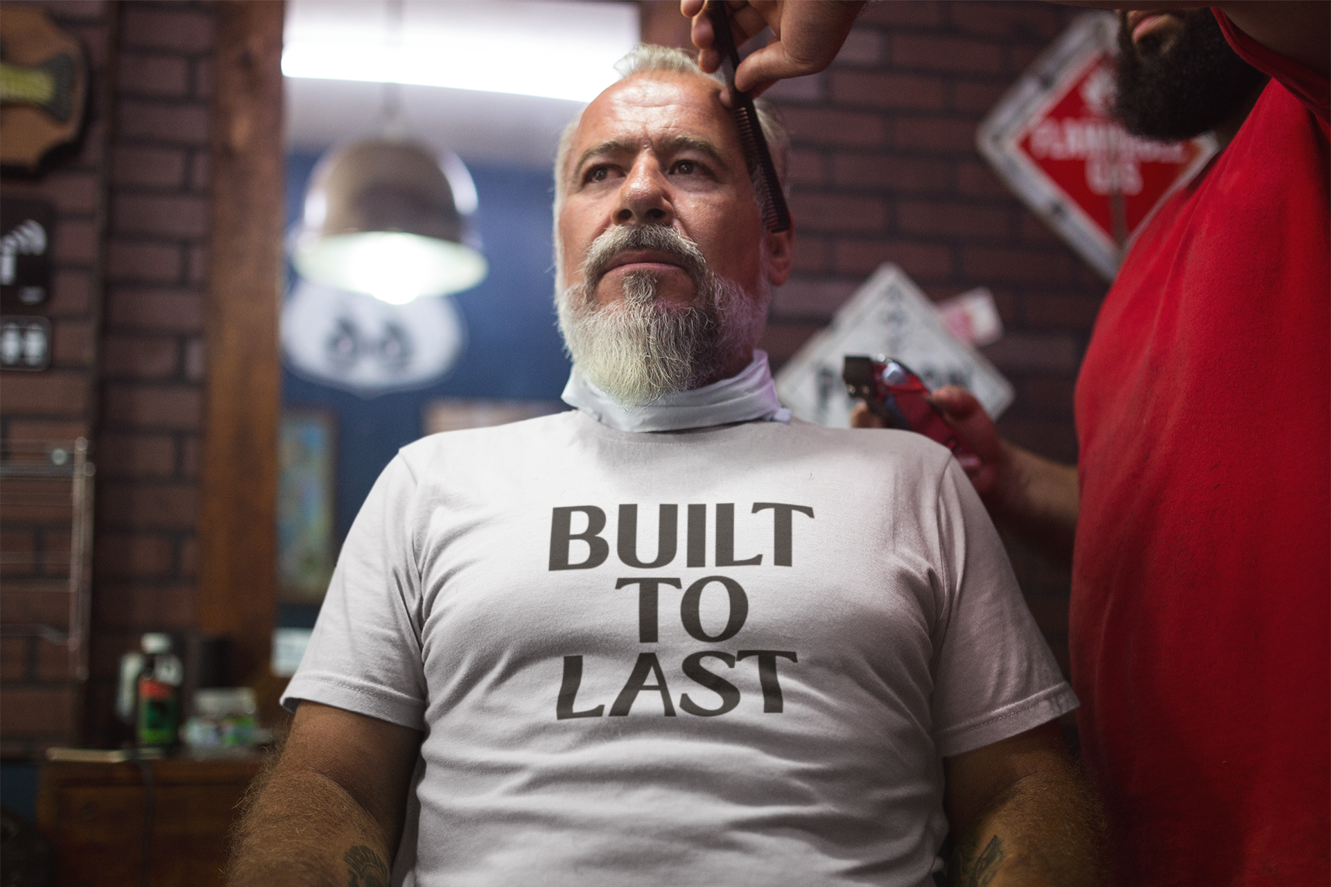 An older man sits confidently in a barbershop chair wearing a white t-shirt that reads “Built to Last,” exuding strength, wisdom, and timeless grit.