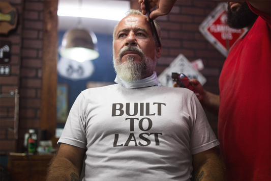 An older man sits confidently in a barbershop chair wearing a white t-shirt that reads “Built to Last,” exuding strength, wisdom, and timeless grit.
