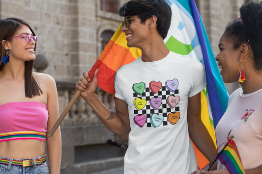 A smiling man at a Pride event holds a rainbow flag while chatting with friends. He wears a white t-shirt featuring colorful candy hearts on a checkerboard background with affirmations like “Queer Joy,” “They/Them,” “Chosen Fam,” and “So Damn Valid.” Two friends in bright rainbow accessories smile beside him, capturing the spirit of joy and solidarity.