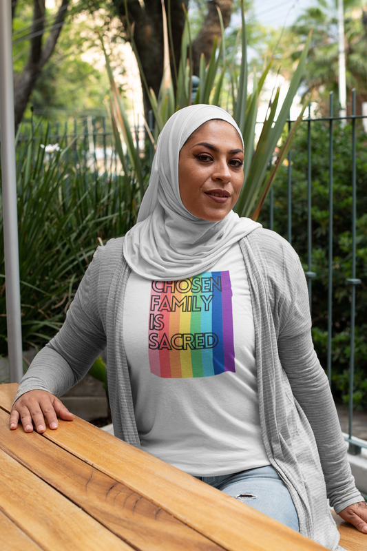 A woman wearing a light gray hijab and cardigan sits confidently at a wooden table outdoors, wearing a white t-shirt that reads “Chosen Family Is Sacred” over a rainbow-striped background. She smiles slightly, exuding pride and warmth.