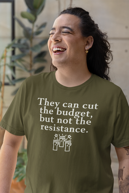 A smiling person with long dark hair and gold earrings wears an olive green t-shirt that reads, “They can cut the budget, but not the resistance,” with an illustration of three raised fists below. They are standing in a sunlit indoor space with plants in the background, exuding joy and resilience.