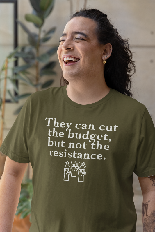 A smiling person with long dark hair and gold earrings wears an olive green t-shirt that reads, “They can cut the budget, but not the resistance,” with an illustration of three raised fists below. They are standing in a sunlit indoor space with plants in the background, exuding joy and resilience.