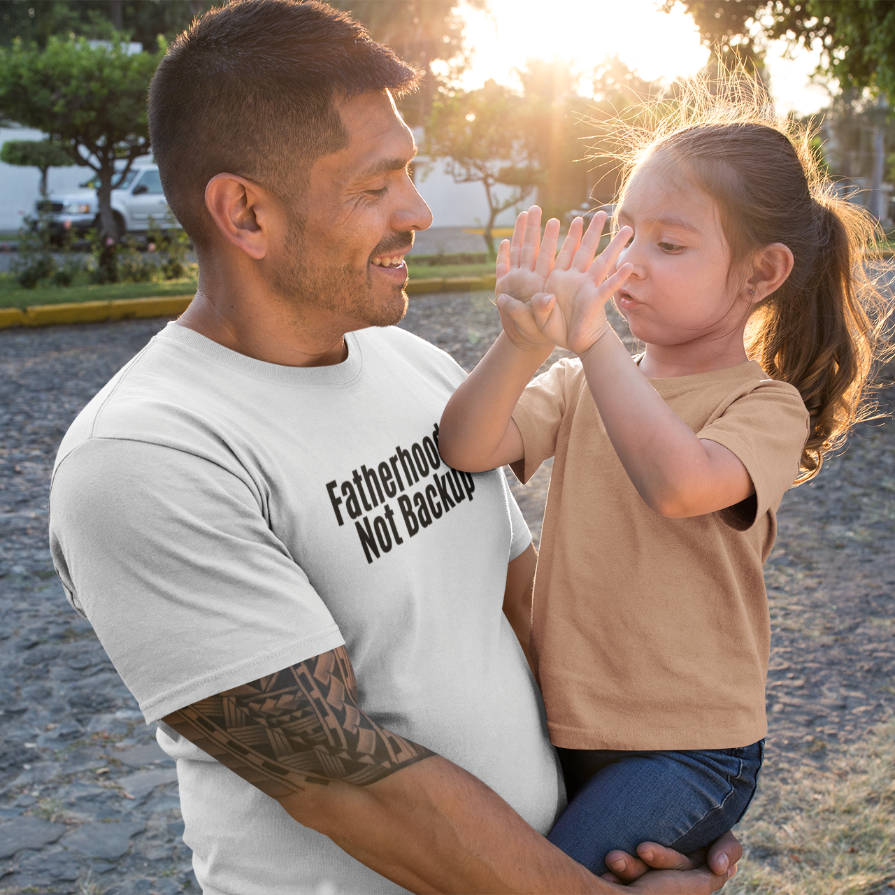 A smiling dad holds his daughter outside while wearing a white "Fatherhood Isn’t Backup" t-shirt by The Unfussy Thread Collective.
