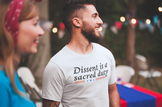 A bearded man laughs with friends at an outdoor party while wearing a white t-shirt that reads “Dissent is a sacred duty” with red and blue graphic lines and stars. The backyard is decorated for a July 4th celebration with string lights and bunting.