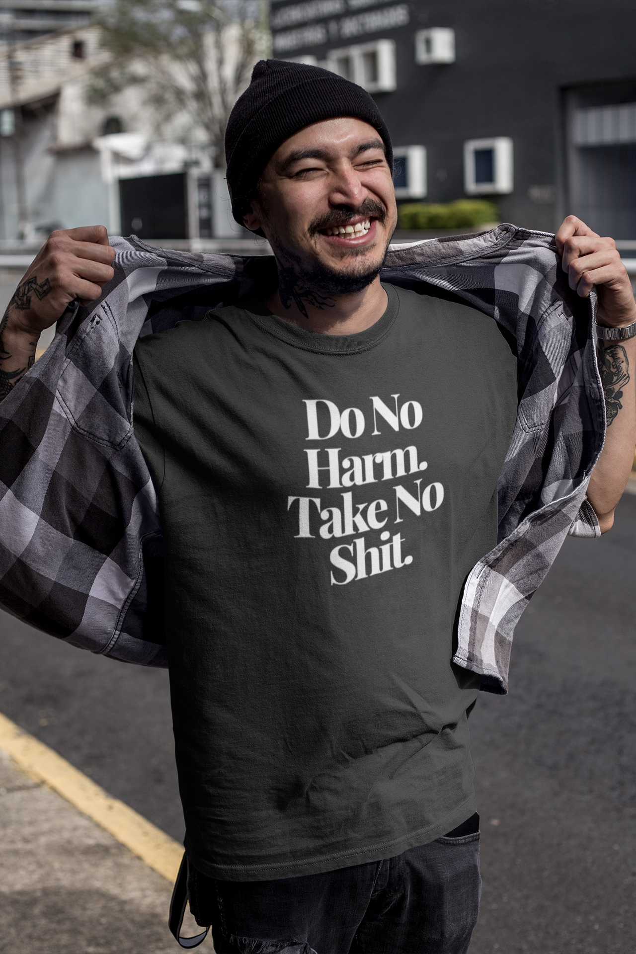 Tattooed smiling man wearing a black beanie and plaid shirt open to reveal a dark “Do No Harm. Take No Shit.” t-shirt, posing confidently on a city street under bright daylight.