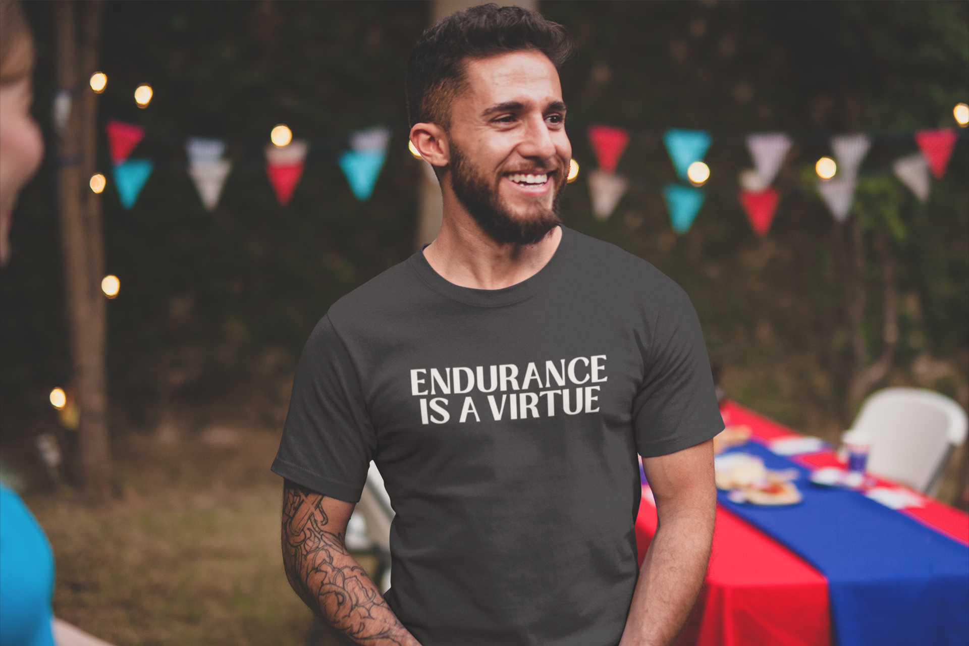 A smiling man at an outdoor gathering wears a dark t-shirt that reads “Endurance Is a Virtue,” standing confidently near a picnic table decorated for a summer event.