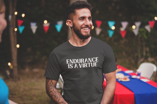 A smiling man at an outdoor gathering wears a dark t-shirt that reads “Endurance Is a Virtue,” standing confidently near a picnic table decorated for a summer event.
