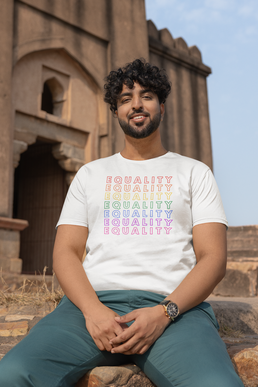 A man sits outside a historic stone building wearing a Classic White “EQUALITY” Pride Gradient t-shirt from The Unfussy Thread Collective.