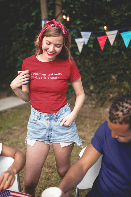 A smiling woman in a red headband and high-waisted denim shorts holds a drink and looks down at her red t-shirt, which reads “Freedom without justice is theater.” She stands at a festive outdoor barbecue with patriotic bunting in the background.
