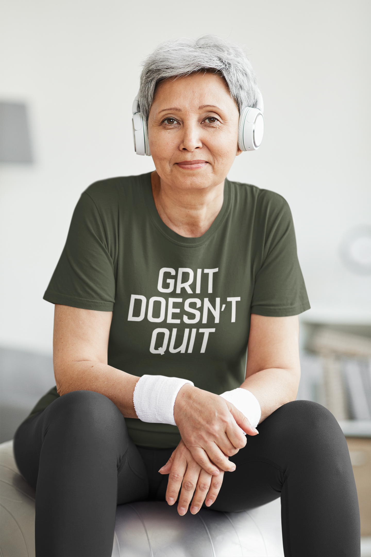 Older woman with short gray hair wearing headphones and a green "Grit Doesn’t Quit" t-shirt, sitting on an exercise ball with wristbands in a fitness space.