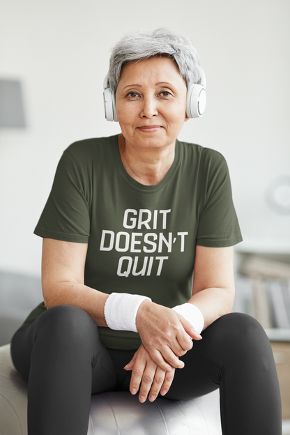 Older woman with short gray hair wearing headphones and a green "Grit Doesn’t Quit" t-shirt, sitting on an exercise ball with wristbands in a fitness space.
