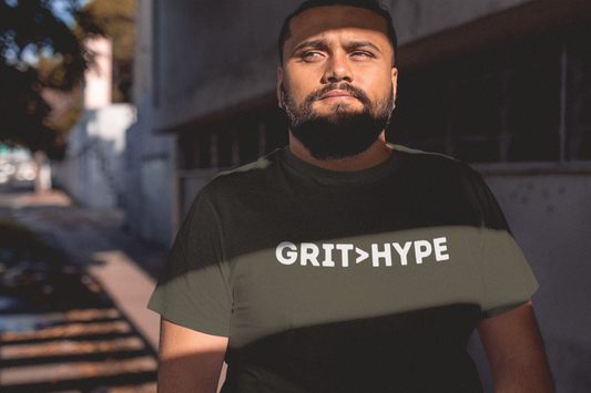 A bearded Latino man stands in dramatic lighting, wearing a dark t-shirt that reads “GRIT > HYPE,” embodying strength, confidence, and quiet defiance.