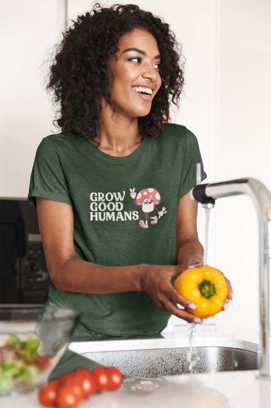A woman wearing a Heather Forest “Grow Good Humans” t-shirt with a peace-sign mushroom graphic while washing vegetables in the kitchen.