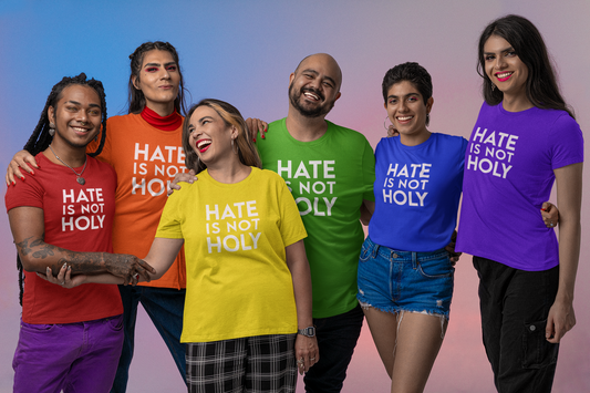 A diverse group of six friends wearing colorful t-shirts in rainbow colors that read “Hate Is Not Holy.” They are smiling, hugging, and standing together against a soft blue and pink gradient backdrop, radiating unity and pride.