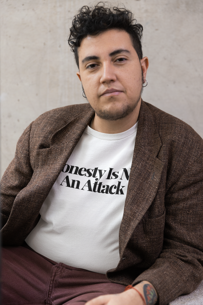 Person with short curly hair wearing a brown textured blazer over a white “Honesty Is Not An Attack” t-shirt, seated against a minimalist concrete wall.